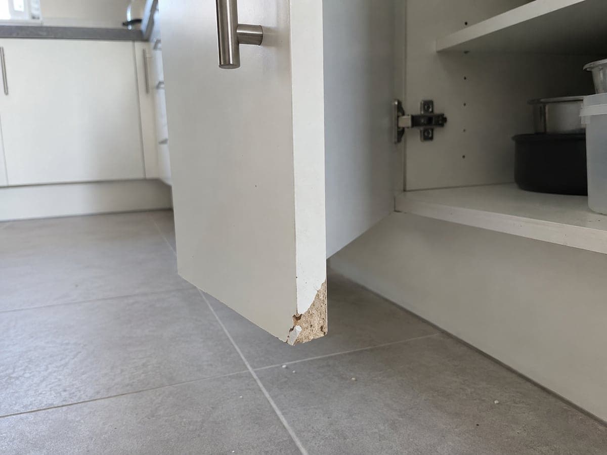 The lower outer corner of the white base-cabinet door is broken away, exposing the particleboard core.